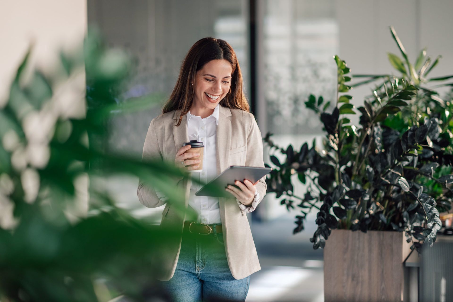 Portrait of business causal businesswoman standing at green corporate office surrounded by plants with tablet in hands and drinking coffee. Close up of manager with coffee smiling at tablet at office.